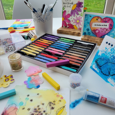 Kreativgarten Klangschalentherapie und kreative Malkurse in Rosenheim. Farbenfroher Kreativtisch mit Pinseln, Papier und bunten Werken – eine Einladung zum freien Gestalten im Kreativgarten von Mea Lumina.